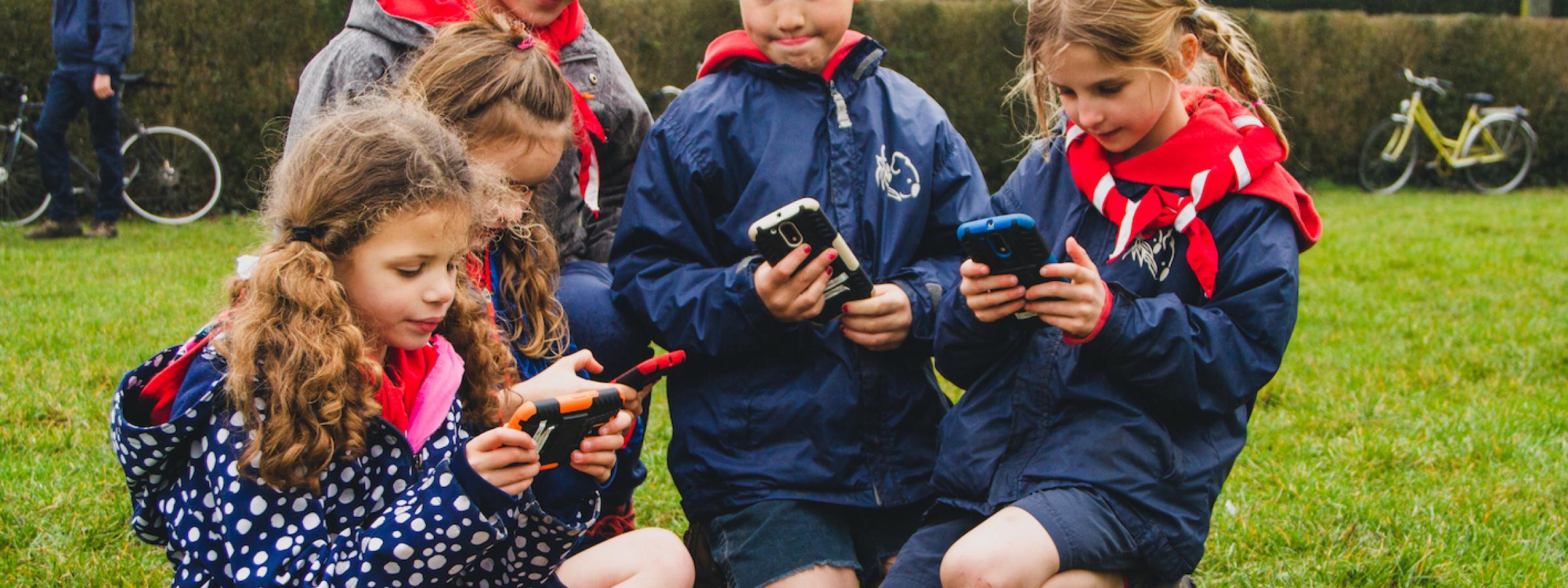 Groepje kinderen van een jeugdbeweging speelt buiten met smartphones
